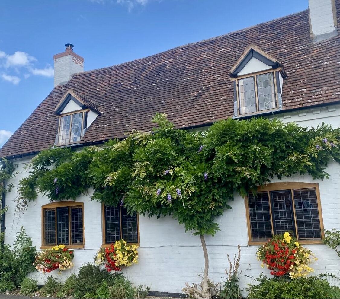 A whitewashed English cottage with wisteria climbing the front, hanging baskets, leaded windows and dormers under a warm summer sky